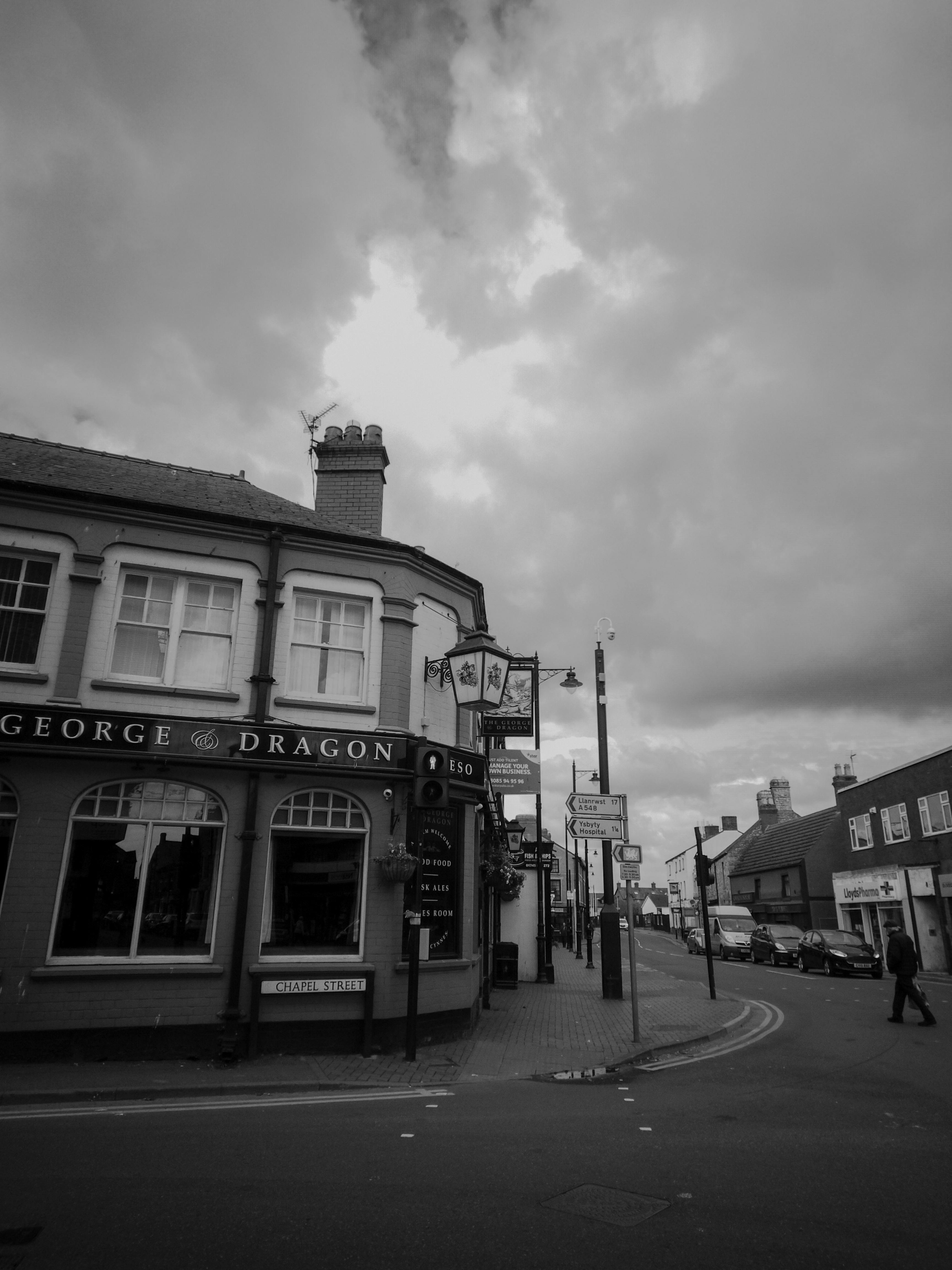 Free A black and white photograph of an urban street corner featuring historic buildings and shops under a cloudy sky. Stock Photo