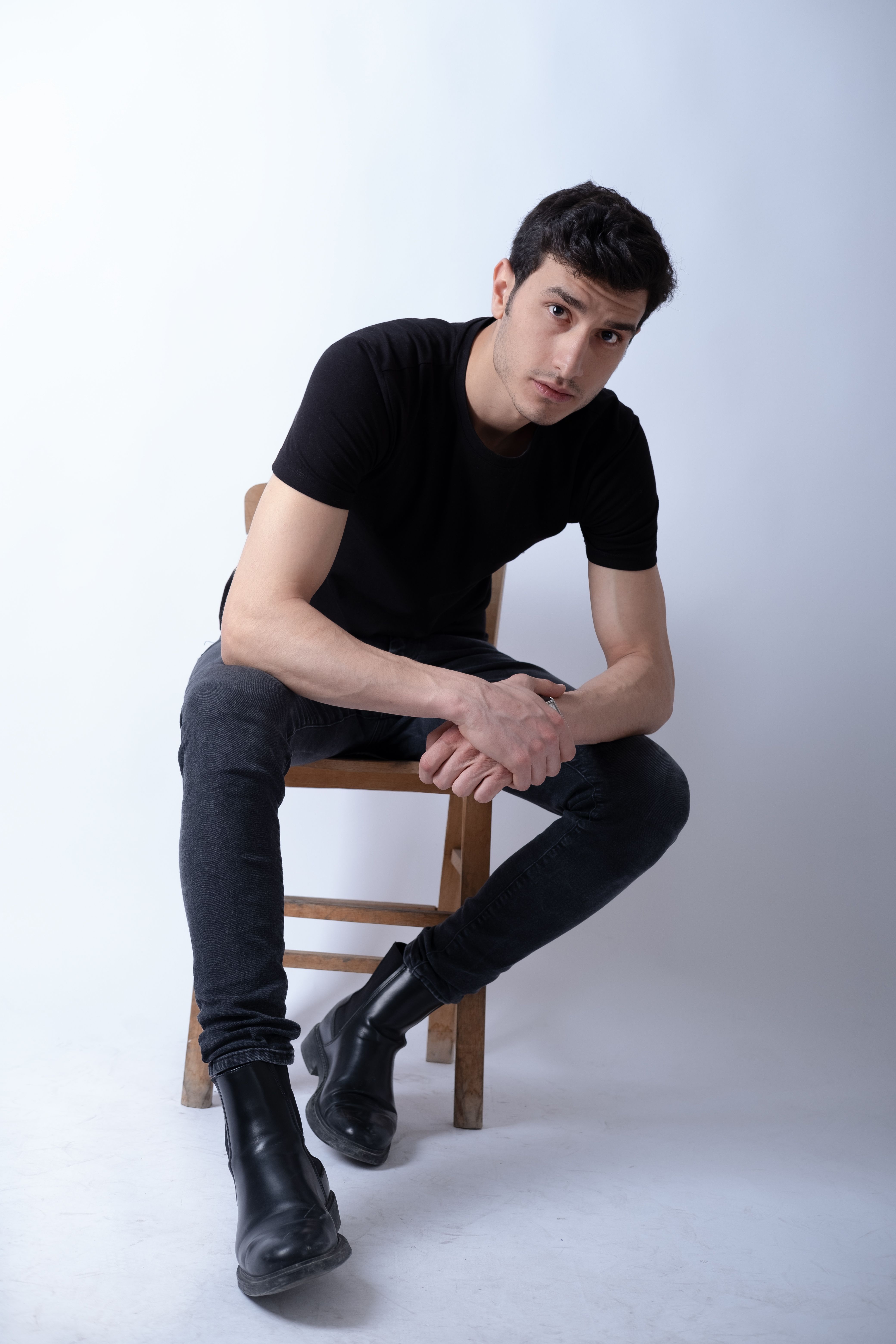 Young man in casual black outfit sitting on a wooden chair against a gray background.