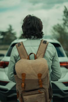 Back view of a traveler with a backpack standing in front of a white car, outdoors.