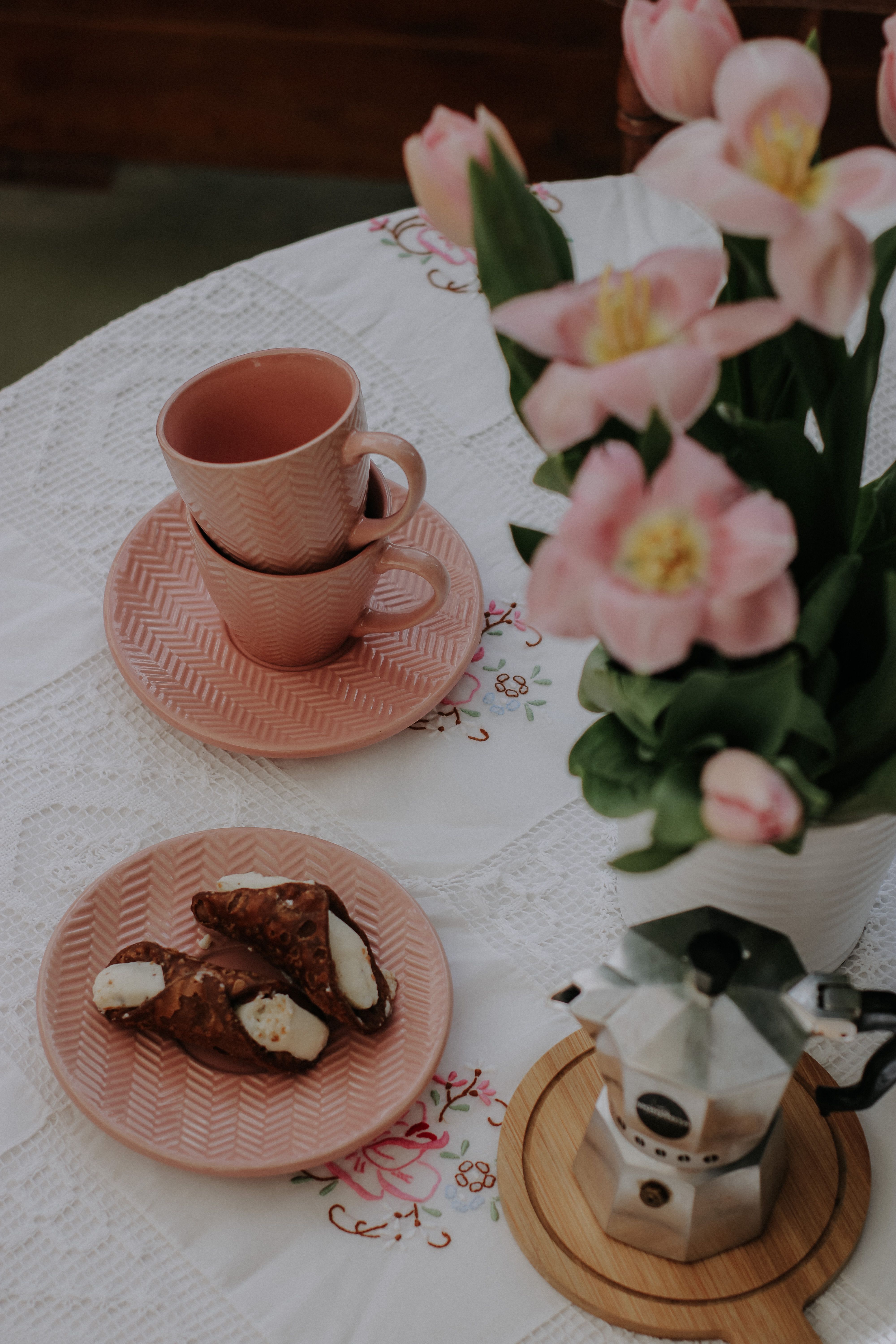 Charming table setting with pink tulips, coffee cups, and sweet pastries on a decorative cloth.