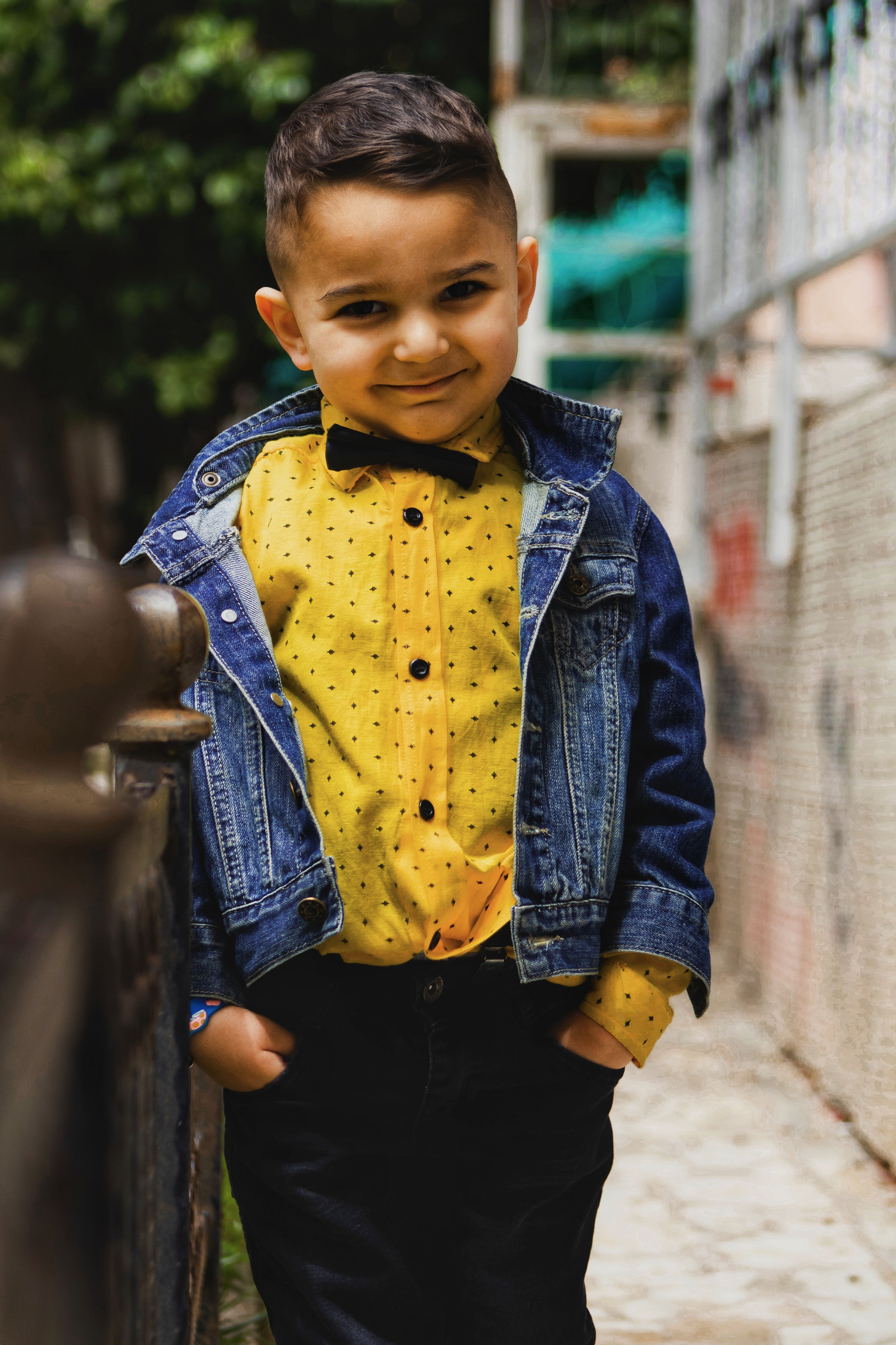 Menino jovem e simpático sorrindo com jaqueta jeans e gravata borboleta, em pé ao ar livre.