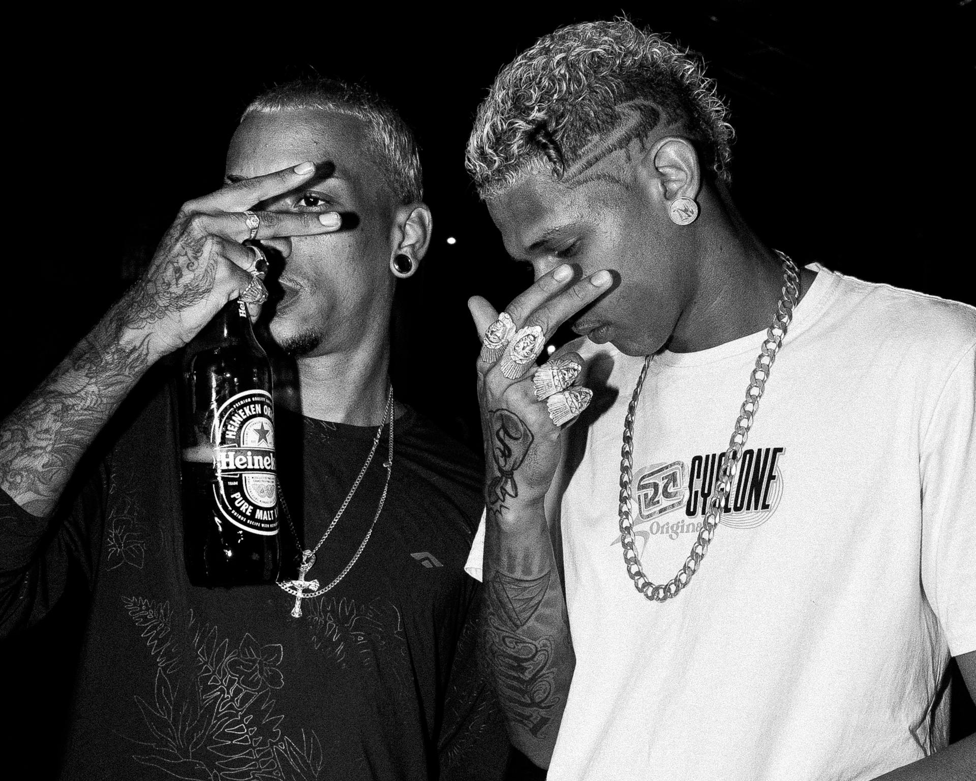 Two stylish men pose with a Heineken bottle, showcasing tattoos and jewelry.