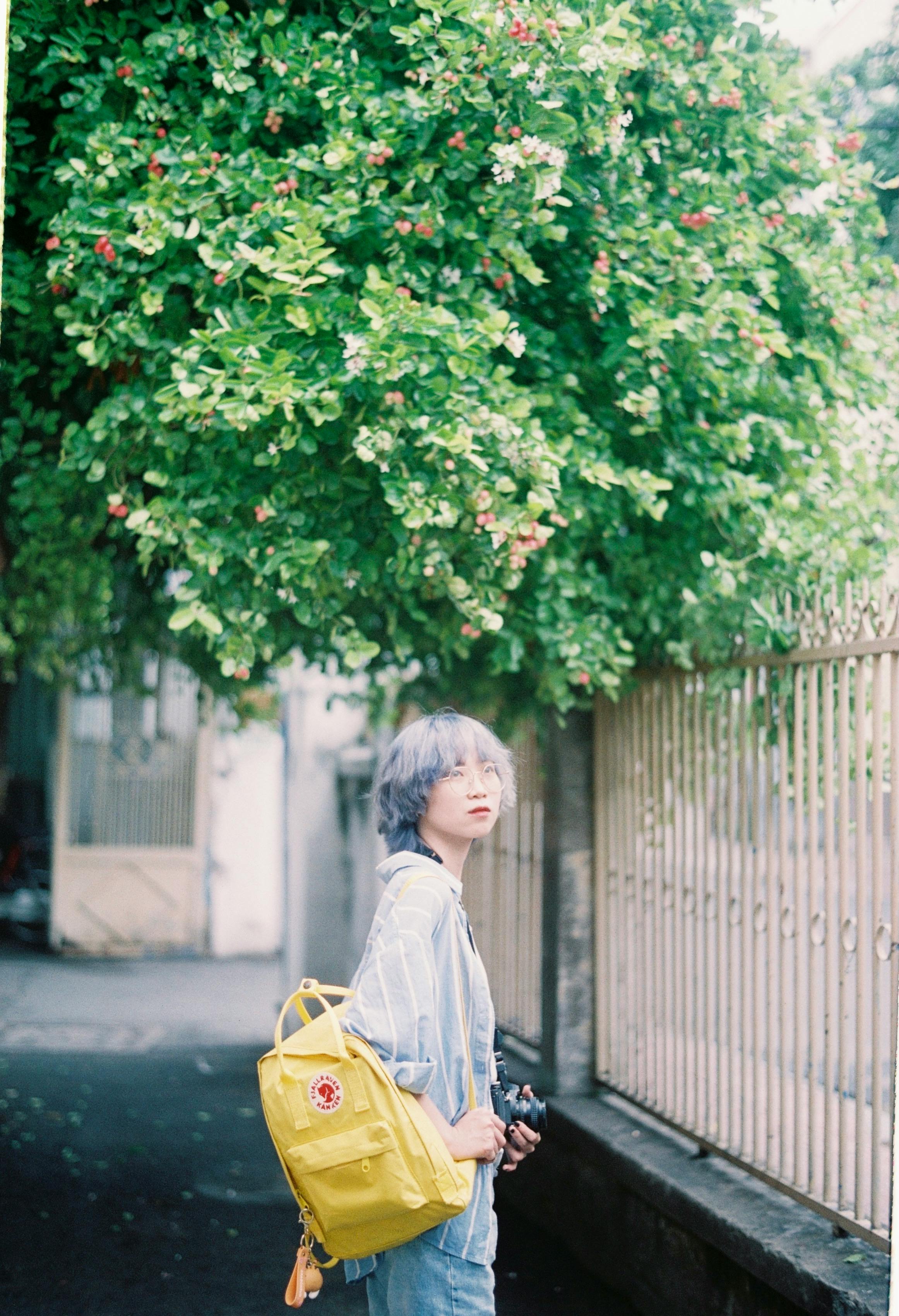 Stylish young woman with purple hair and yellow backpack walking along a leafy sidewalk.