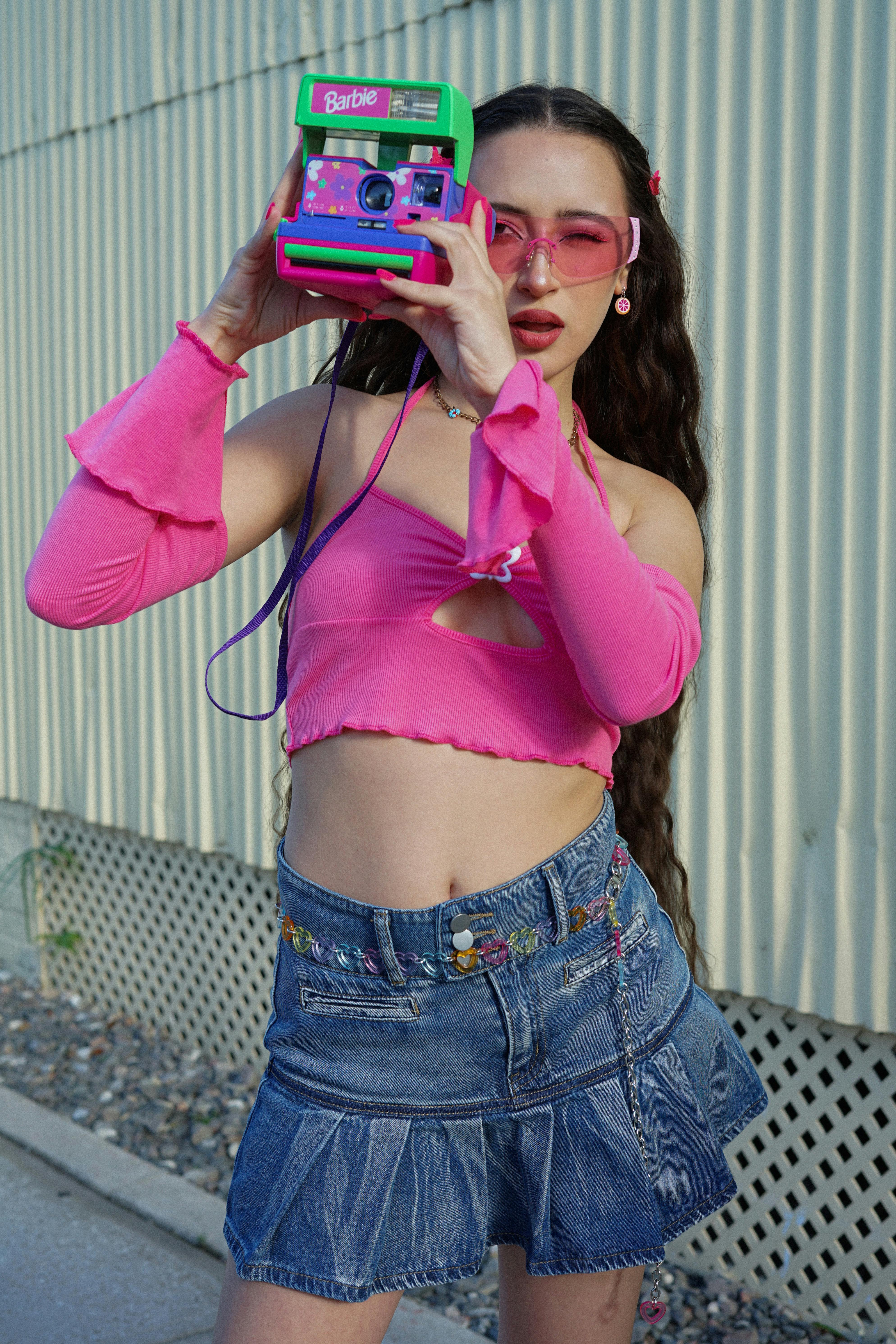 Stylish woman in pink top and denim skirt posing with a colorful polaroid camera outdoors.