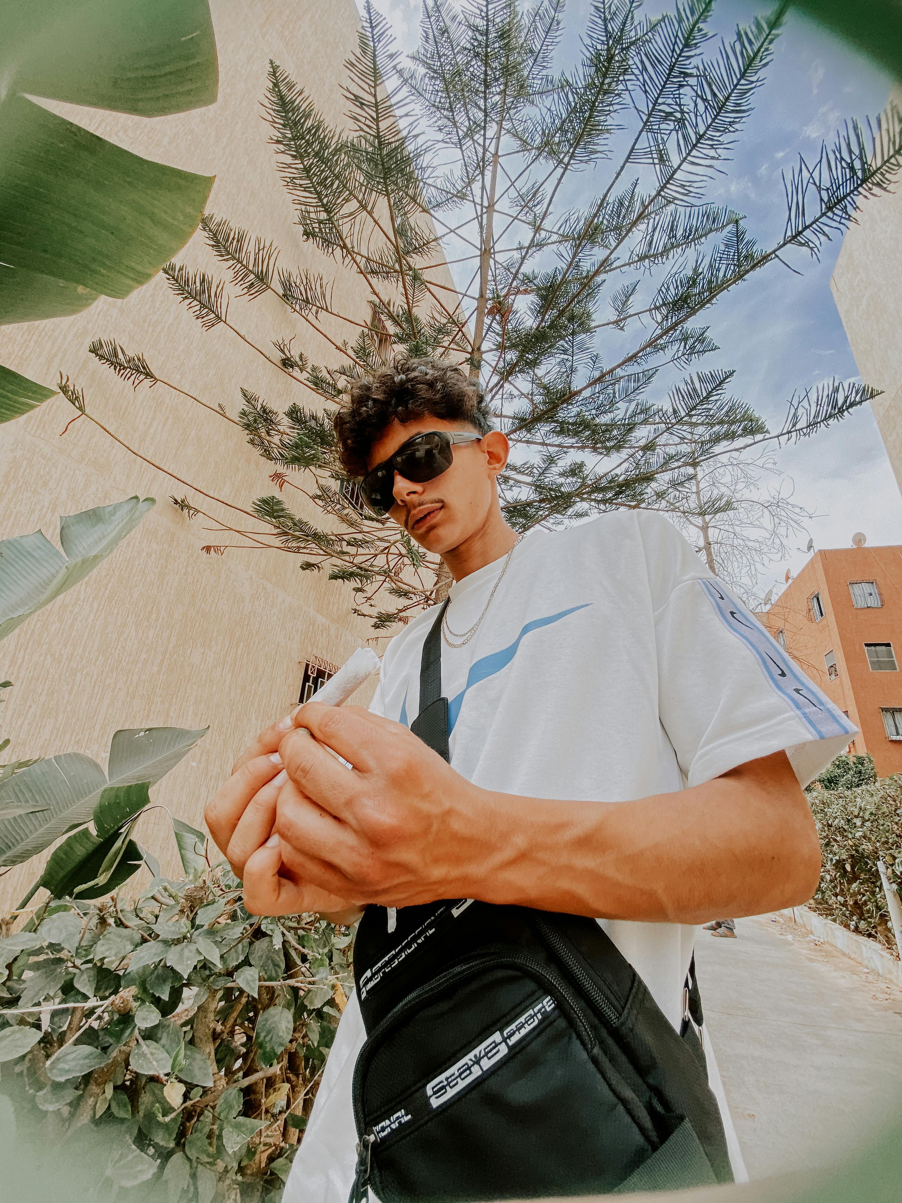 Young man in sunglasses rolls a cigarette outdoors with modern buildings and greenery.