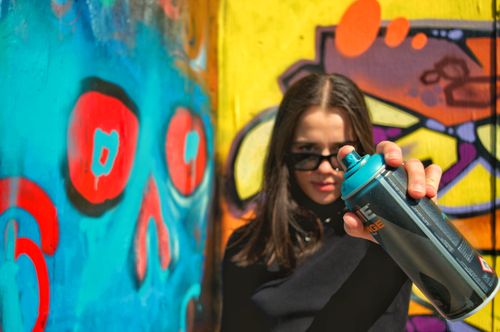 Free Young woman with sunglasses holding spray can in front of colorful graffiti wall outdoors. Stock Photo