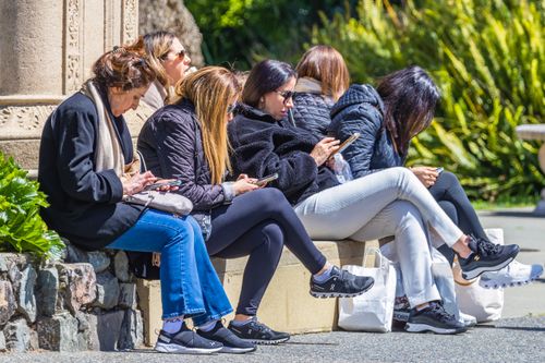 Free Five women sitting outdoors, using smartphones, enjoying sunny day in park. Stock Photo