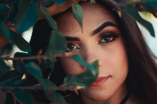Striking close-up portrait of a woman with vivid makeup peering through green foliage.