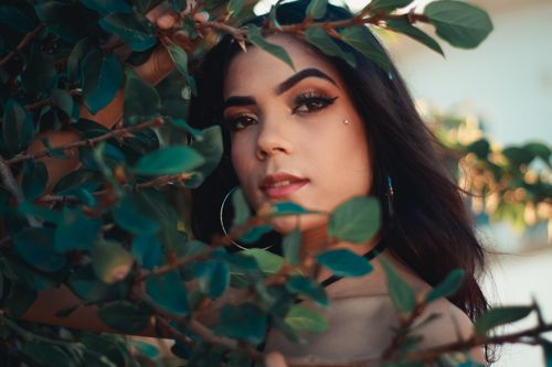 Free Close-up of a woman with makeup posing through lush green branches in natural light. Stock Photo