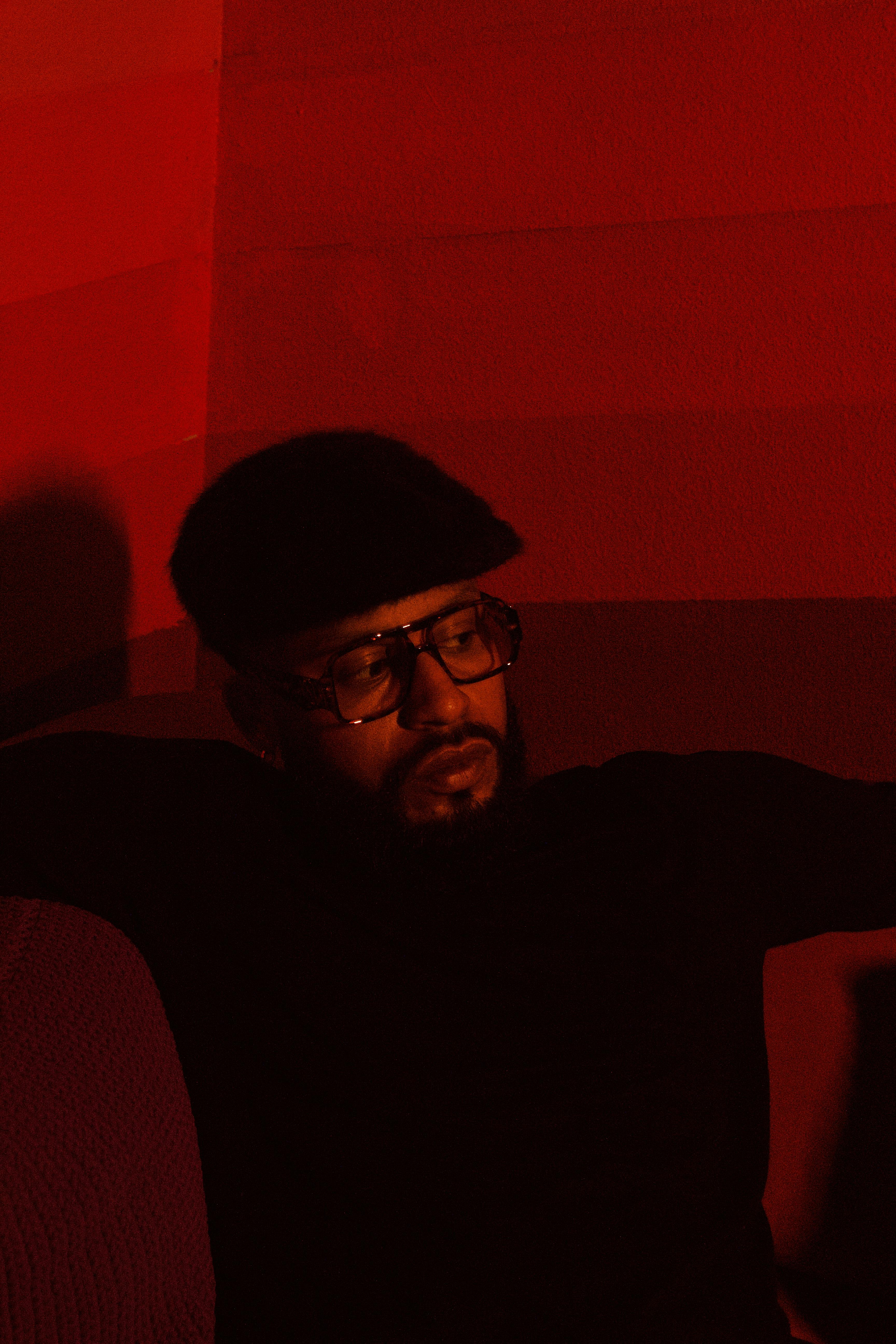 A man in a black sweater and flat cap relaxes in a red-lit room, conveying a moody atmosphere.