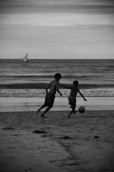 Two boys play football on a beach, captured in black and white.