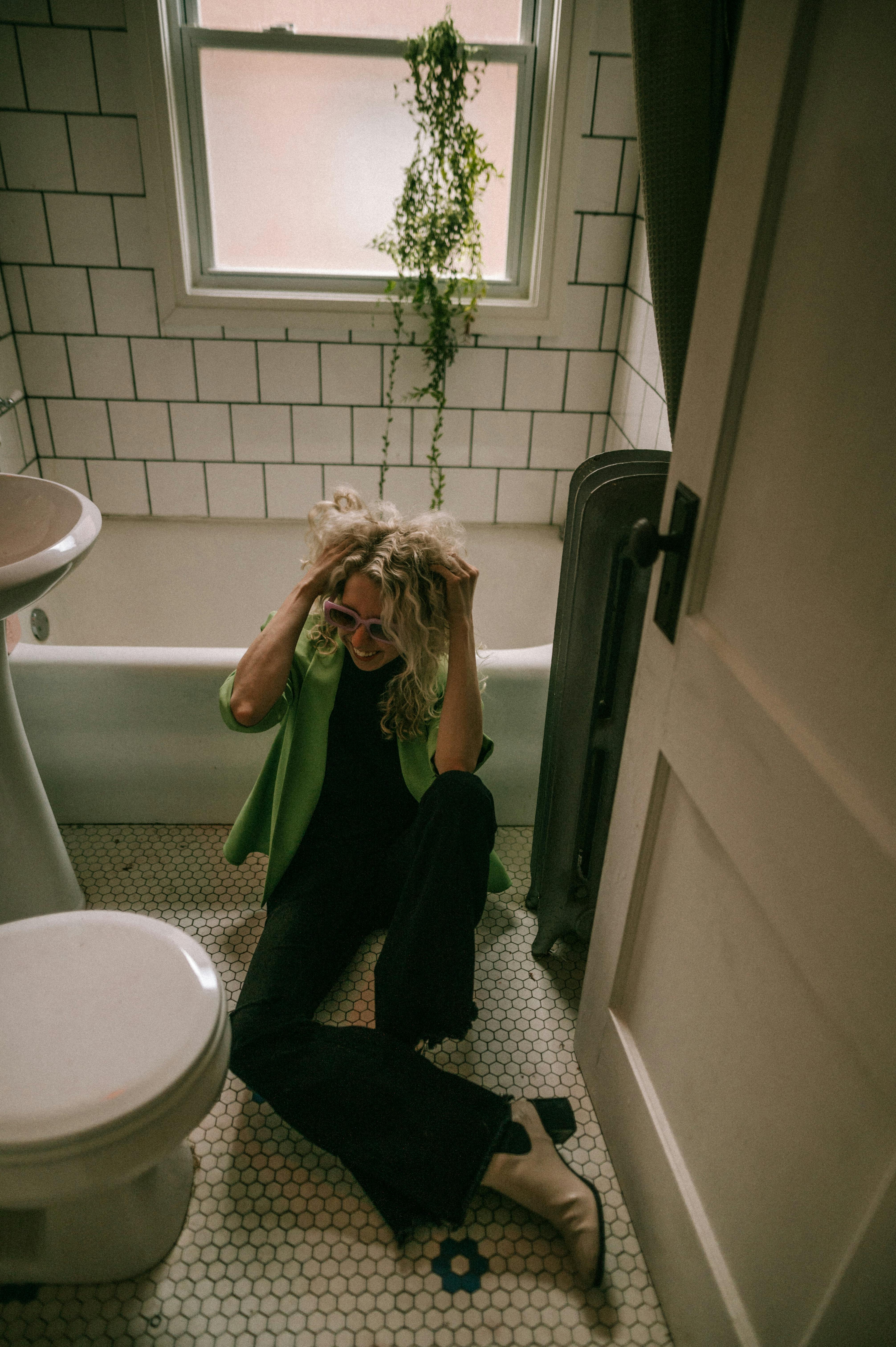 Woman sitting on bathroom floor, wearing a green jacket, in a vintage-style bathroom.