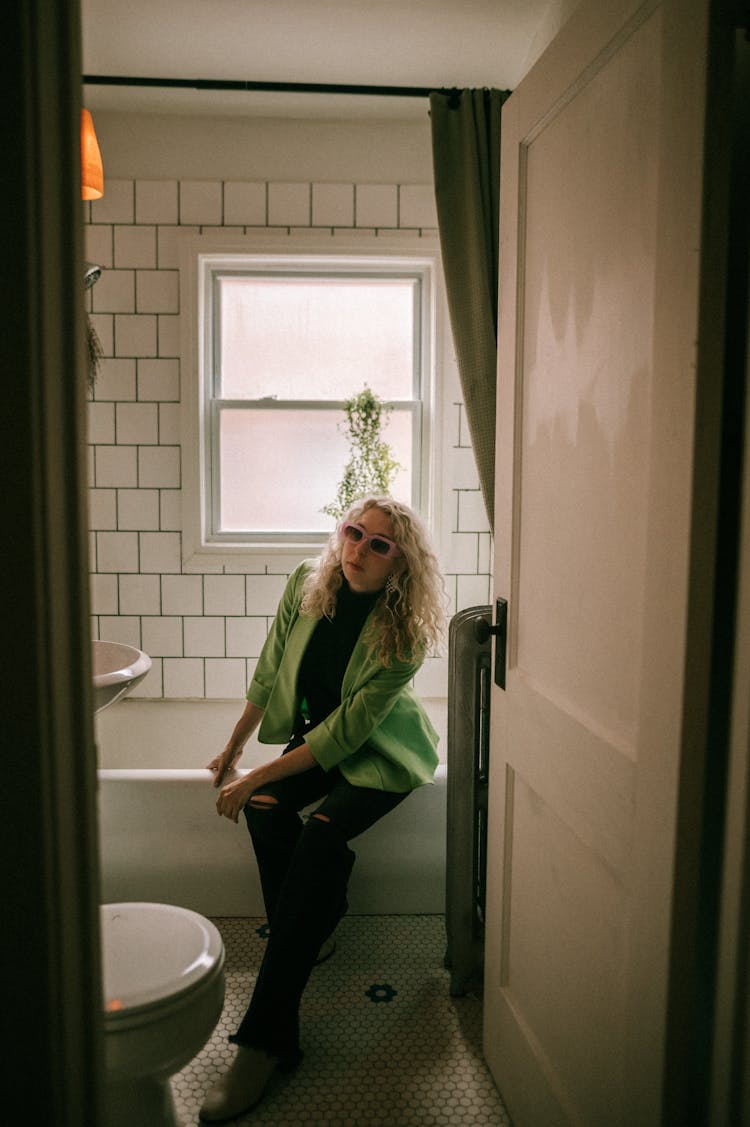 Blonde Woman In Green Jacket Sitting On Bathtub