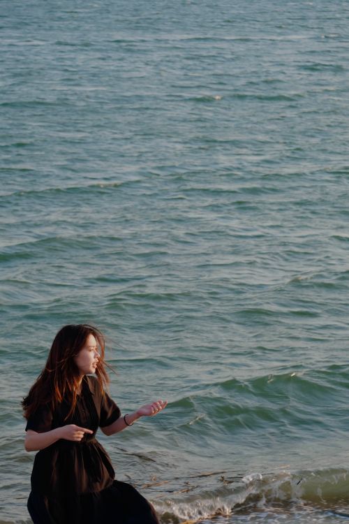 Free Serene woman in a black dress enjoys a peaceful moment by the seaside during sunset. Stock Photo