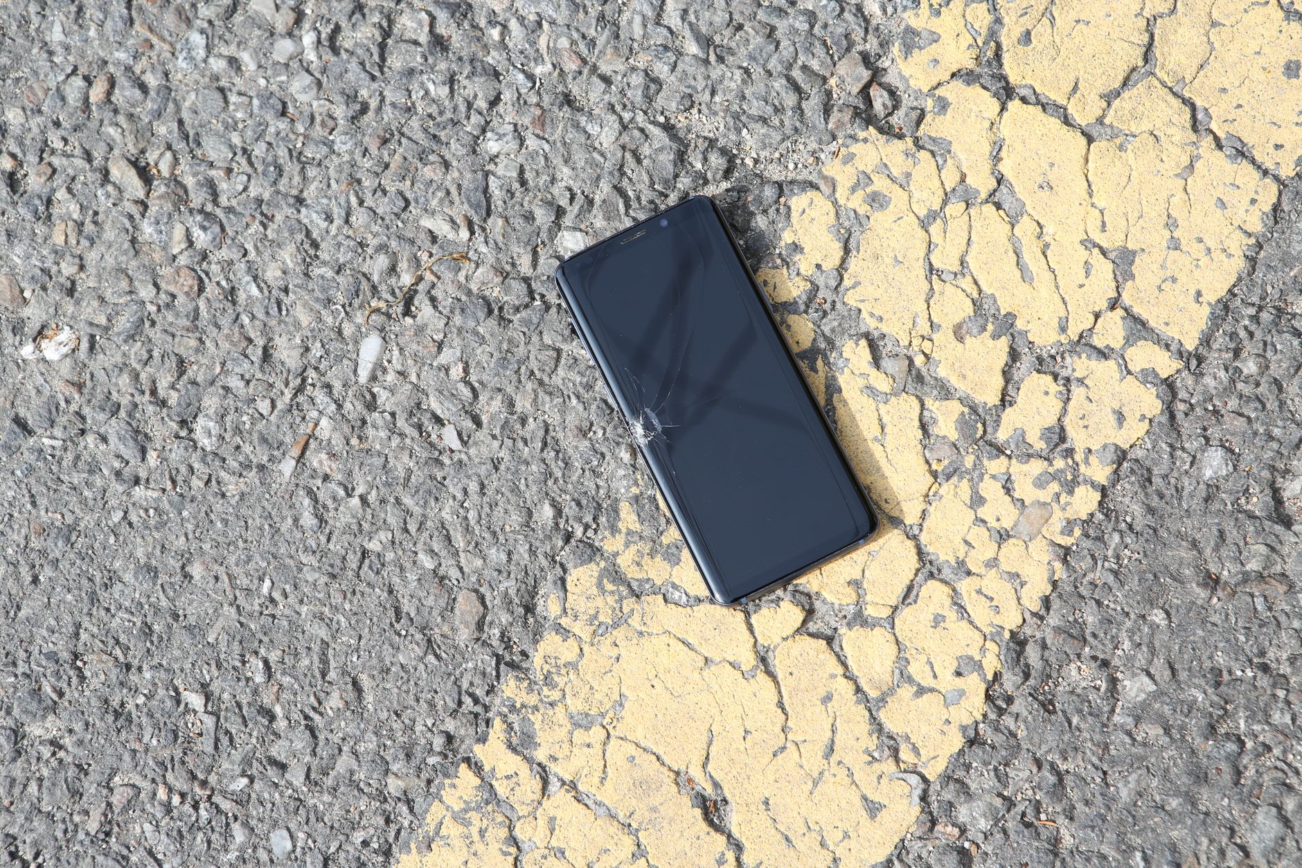 A close-up view of a cracked smartphone lying on a sunlit asphalt road.