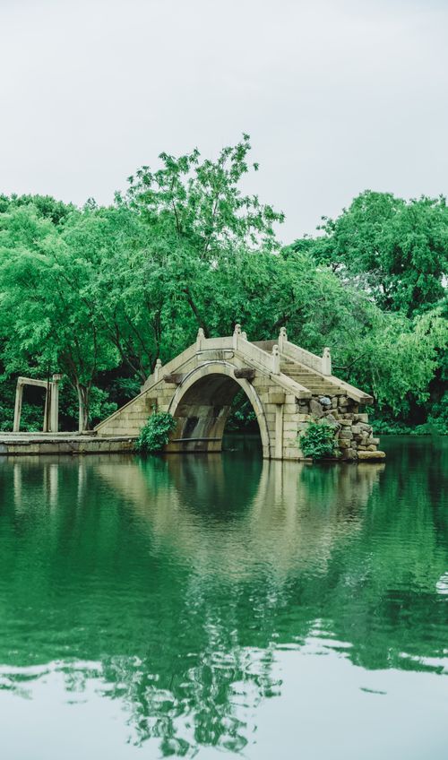Free Tranquil scene of a stone bridge reflected in a calm green pond surrounded by lush trees. Stock Photo