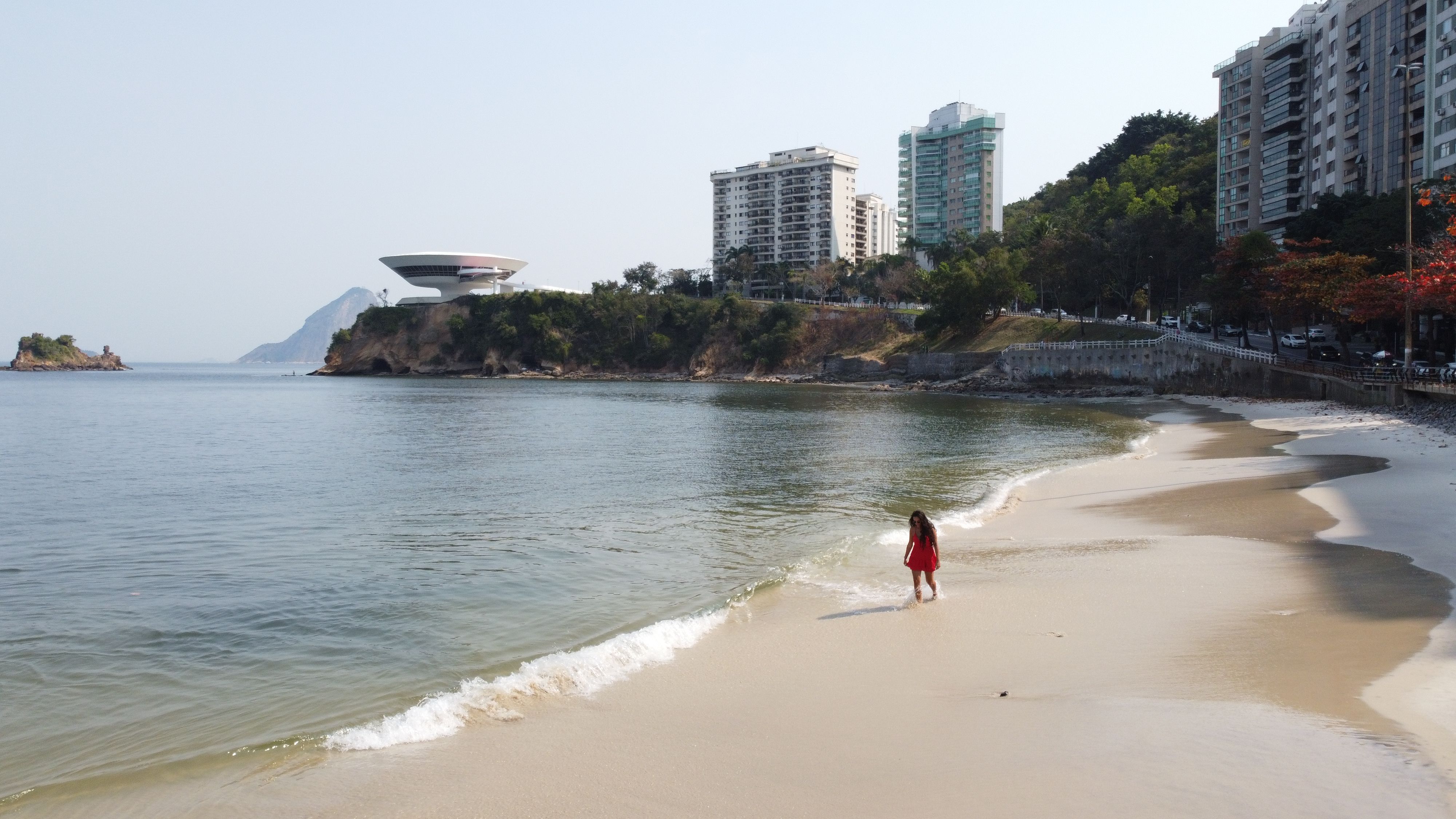 A woman in red strolls on a beach in Niterói, Brazil, with the Museum of Contemporary Art visible in the background.