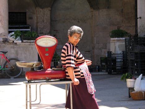 A senior woman stands near a vintage weighing scale in an outdoor marketplace.