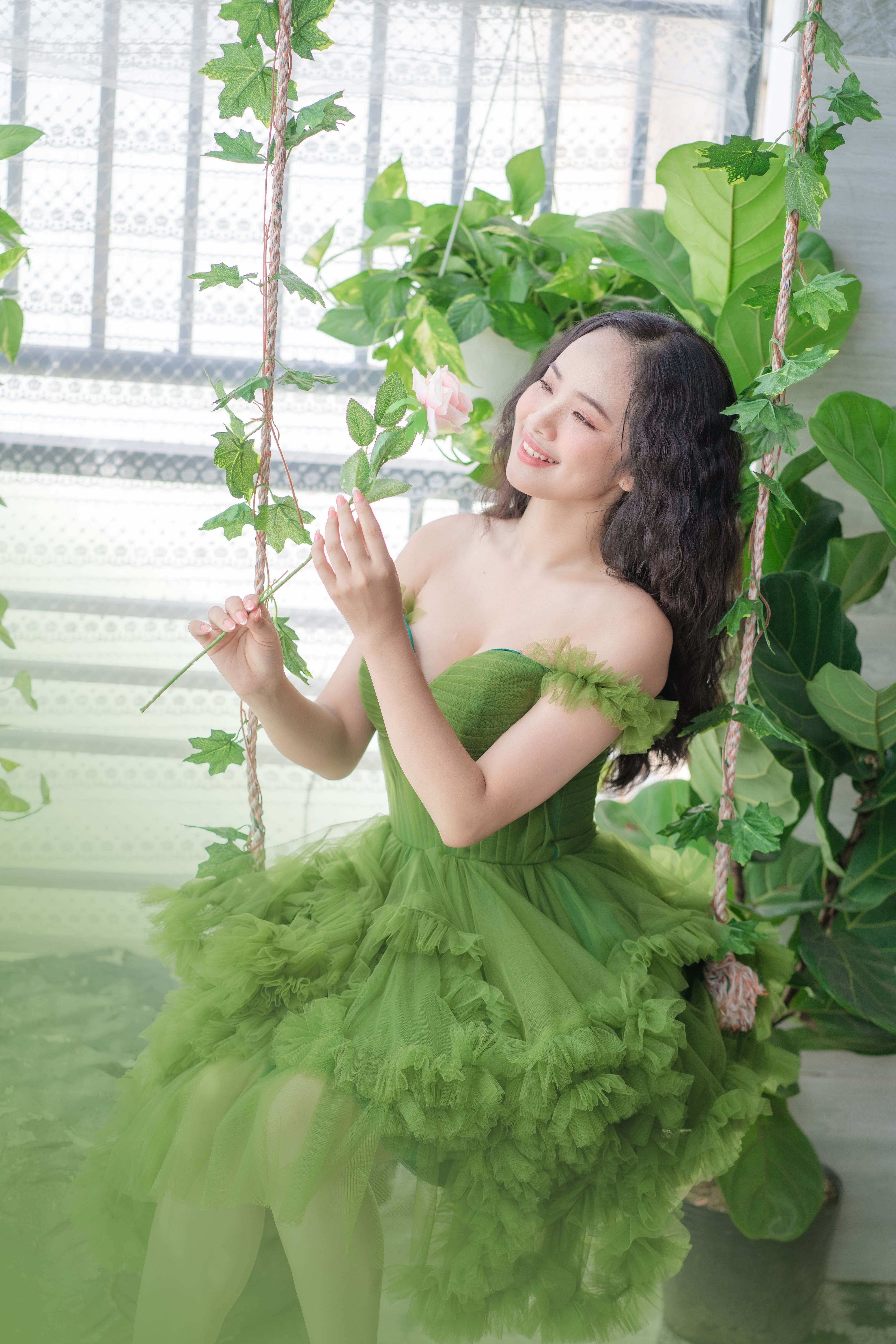 A joyful woman in a green dress enjoying a swing surrounded by lush greenery indoors.