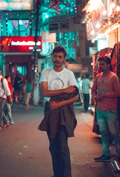 A fashionable man walking through a vibrant, illuminated street market in India at night.