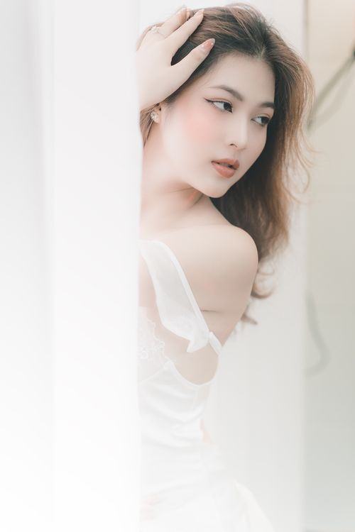 Free Beautiful young Asian woman posing delicately in a soft white dress indoors. Stock Photo