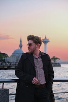 Young man in sunglasses and coat by sea at sunset, with mosque and minaret in the background.