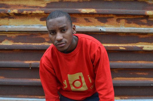 A man in a red pullover posing outdoors against a rustic metal background.