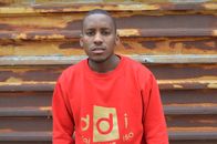Casually dressed young man standing in front of a rusty wall wearing a red sweatshirt.