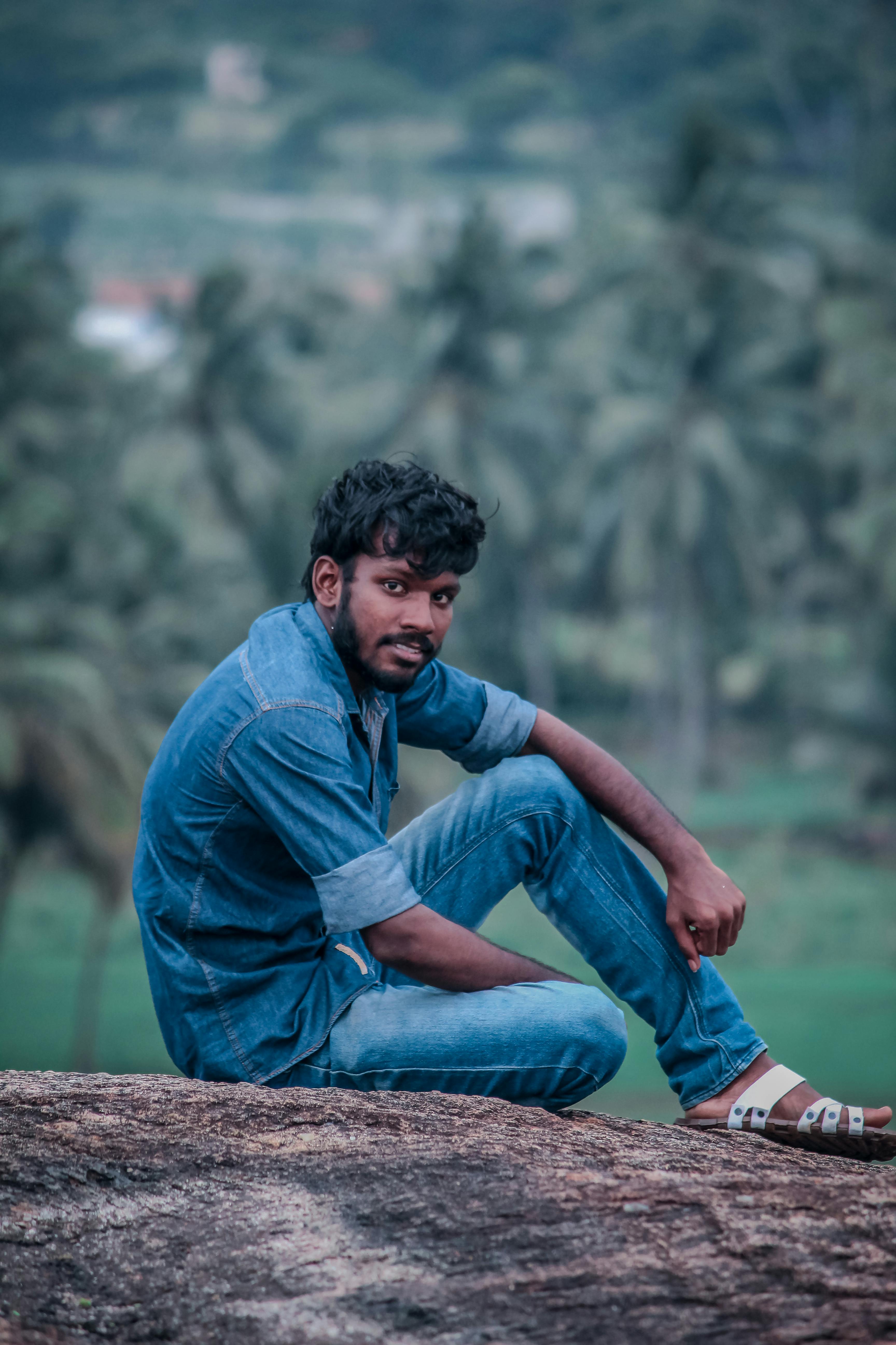 Casual portrait of a young man in denim clothing sitting on a rock with tropical background.