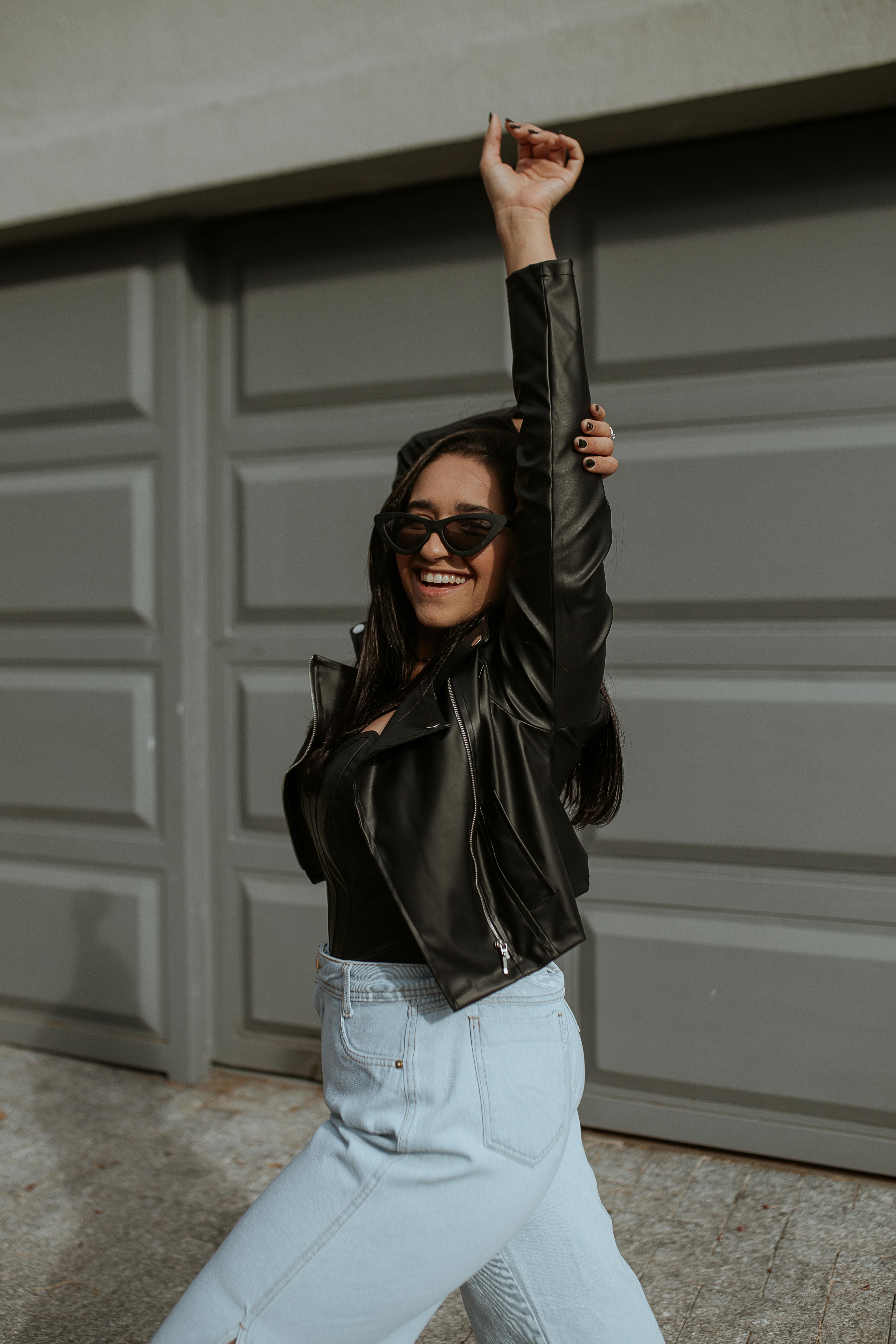 Smiling woman in a leather jacket and sunglasses striking a playful pose outdoors.