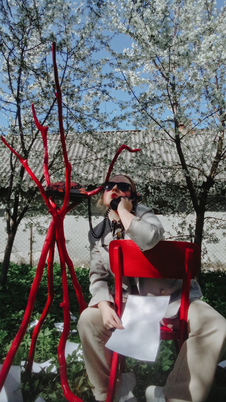 Young Creative Woman Straddling Red Wooden Chair And Talking Over Analog Telephone In Spring Garden