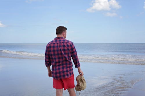 免费 男子手持棕色帽子面对海滩 素材图片