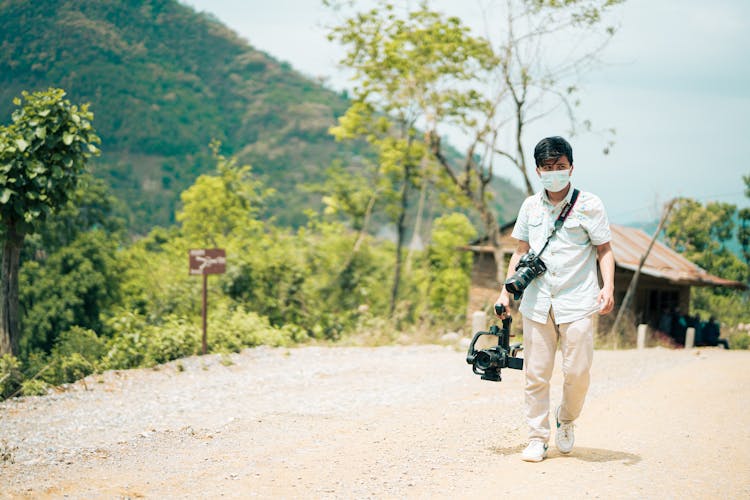 Man In Shirt And With Cameras Walking On Dirt Road In Village