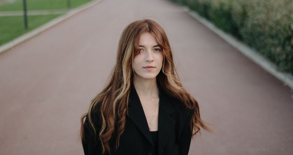 Portrait of a young woman with long hair standing on an outdoor pathway wearing a black jacket.