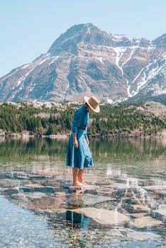 A woman in a blue dress and hat walking on clear rocky shoreline with a mountain backdrop.