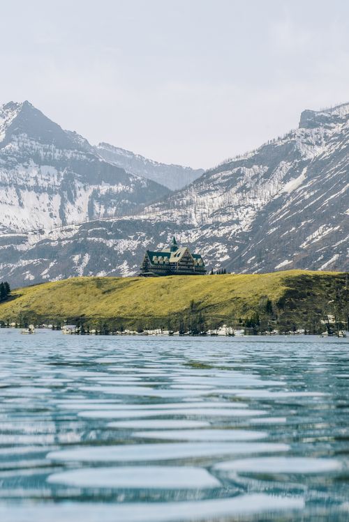 Free A picturesque mountain lodge sits serenely beside a tranquil lake, framed by majestic snow-capped peaks. Stock Photo