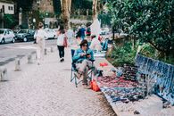 A woman selling handmade jewelry on a sidewalk in a lively urban park, engaging with tourists.