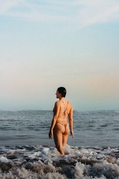 A woman wearing a bikini stands at the shore enjoying the ocean and sunset.