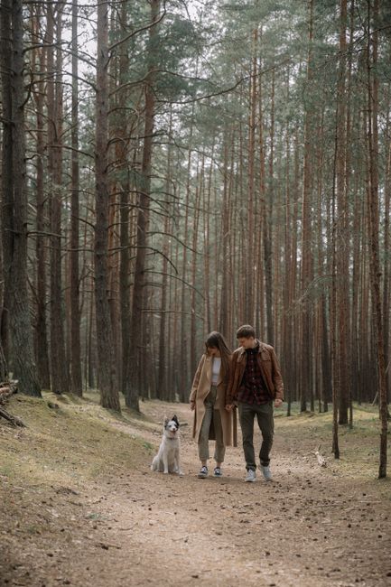A couple enjoys a peaceful walk with their dog along a forest path, surrounded by tall trees.