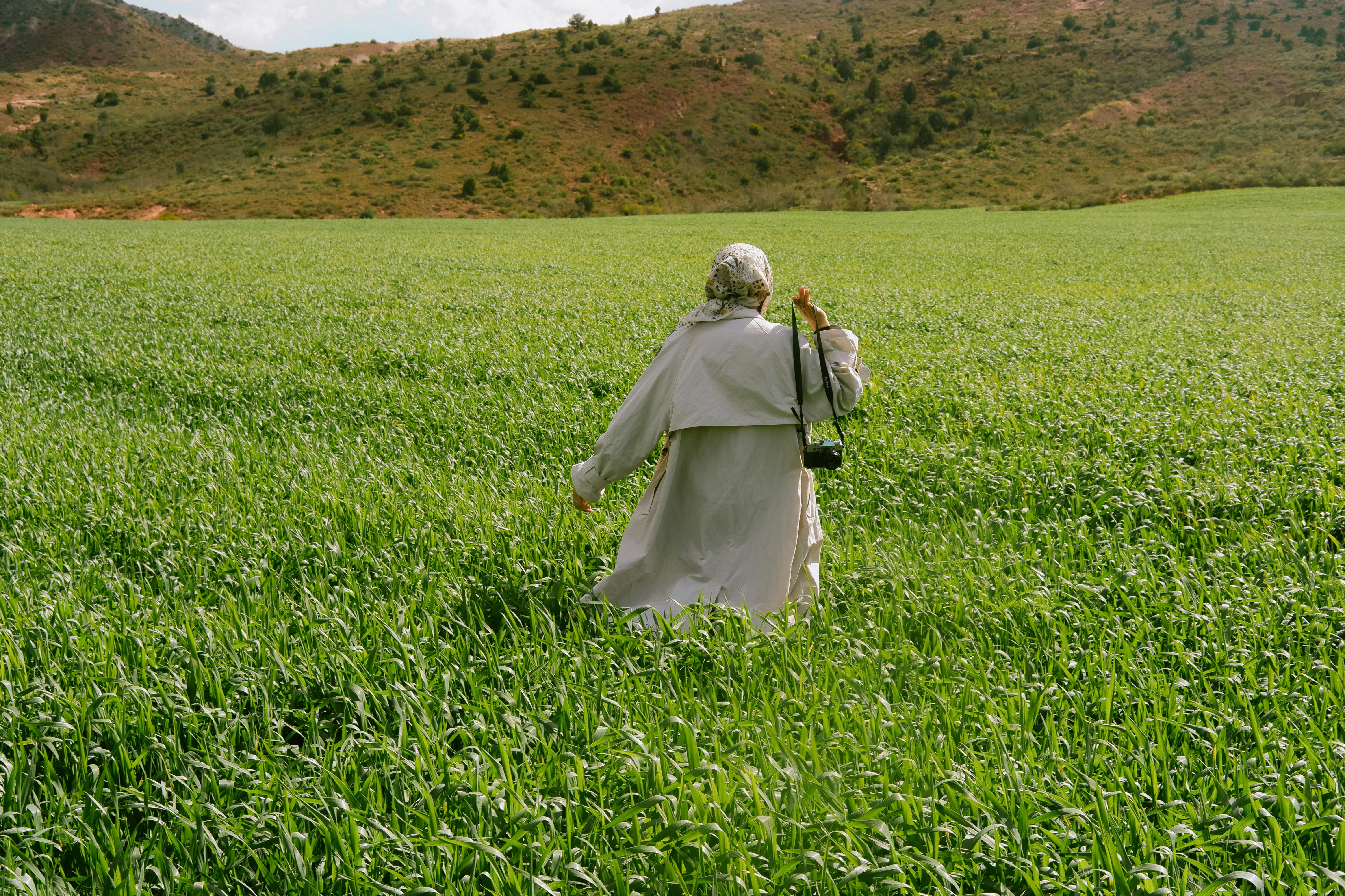 A woman wearing a trench coat and headscarf walks through a lush green field, evoking a sense of freedom.