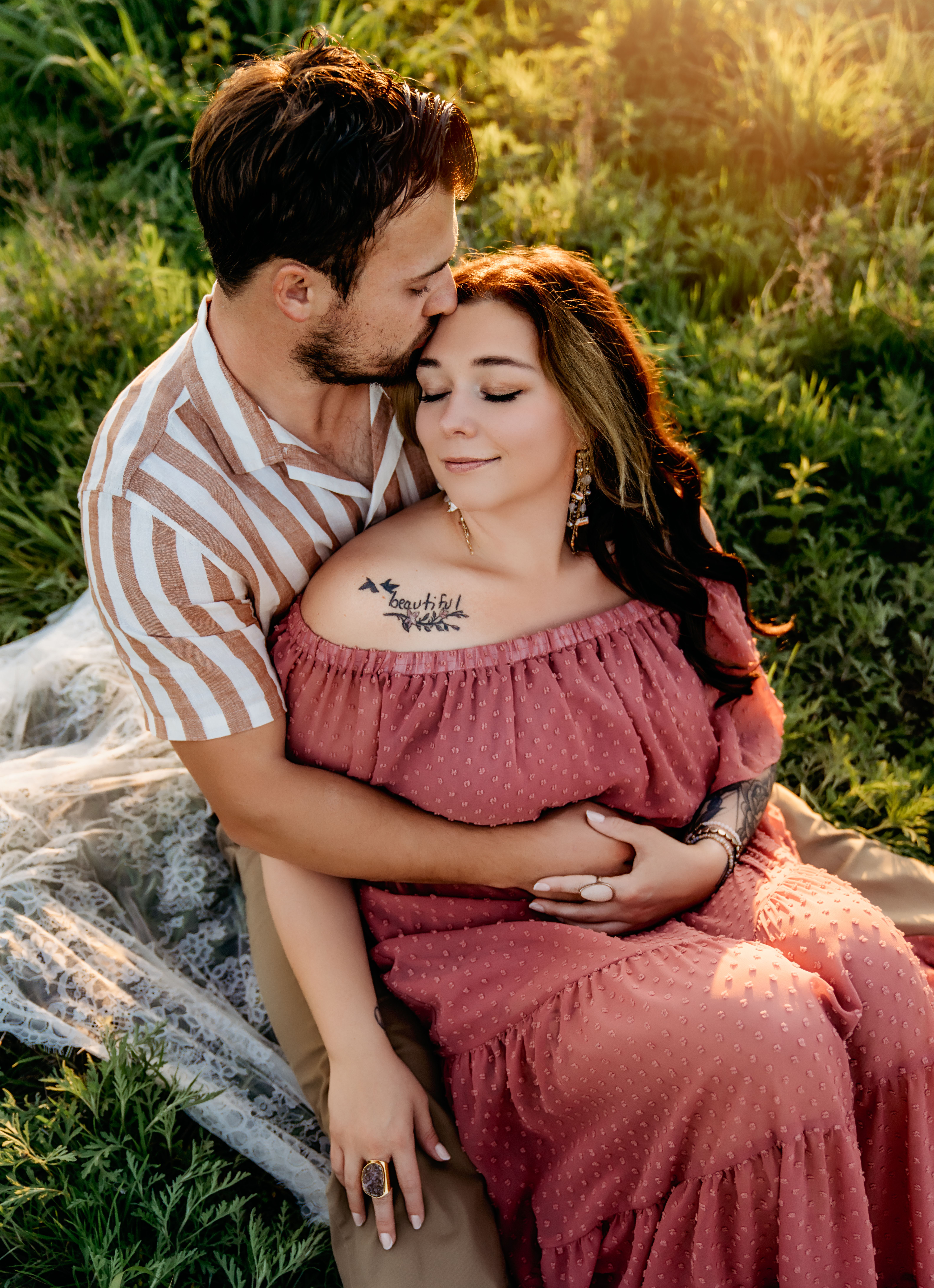 Tender moment of a couple embracing in a sunny field, featuring tattoos and warm lighting.