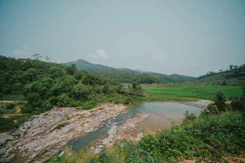 Free A picturesque view of a footbridge spanning a serene river with lush mountains in the background. Stock Photo