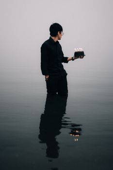 A man in black holding a birthday cake with lit candles, standing in misty waters.