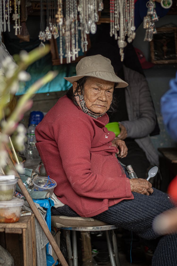 Photo Of An Elderly Woman Sitting On A Chair At A Market Stall 