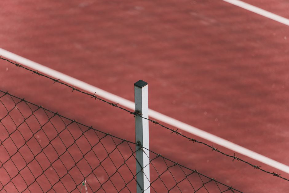 Close-up of a barbed wire fence with a red tennis court in the background, emphasizing urban contrasts.