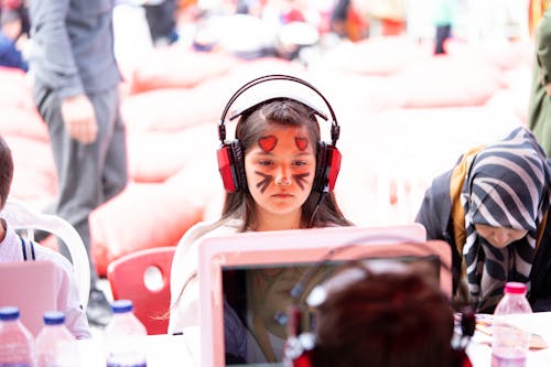 Small Girl with a Painted Face Sitting in Front of a Laptop in Headphones 