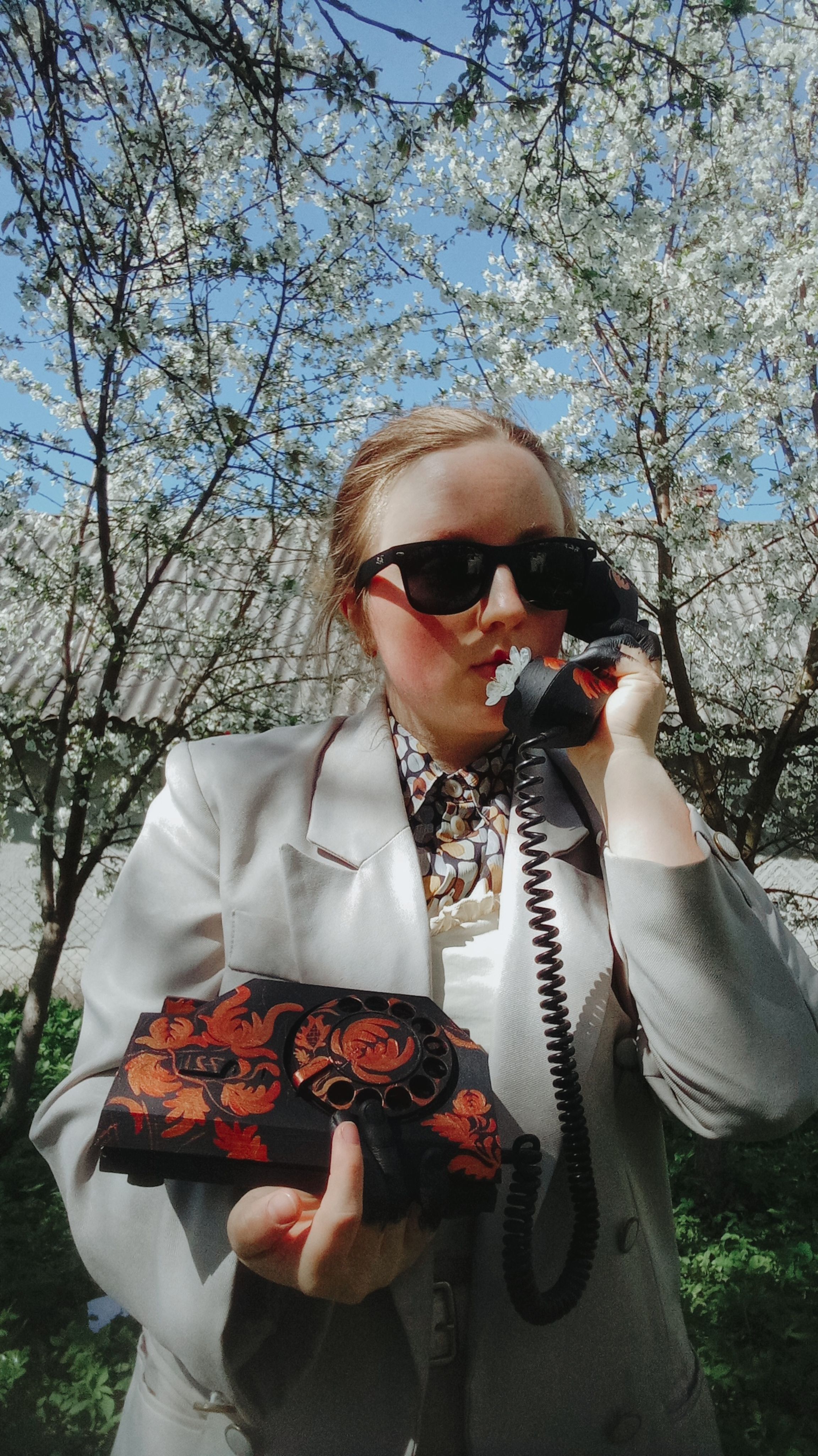 Stylish woman in sunglasses poses outdoors holding a vintage phone against a spring backdrop.