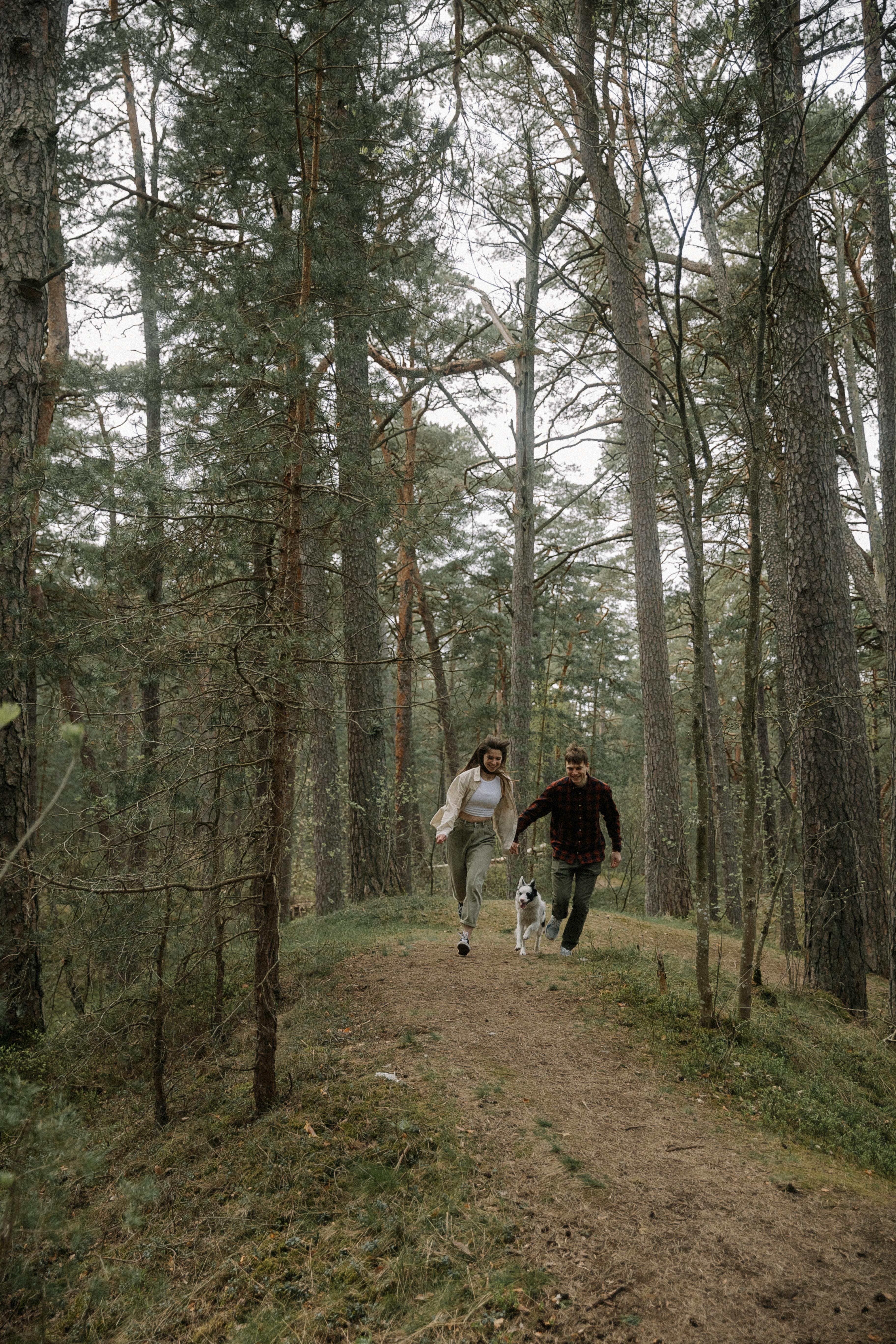 A couple runs joyfully with their dog along a forest pathway, surrounded by tall trees.