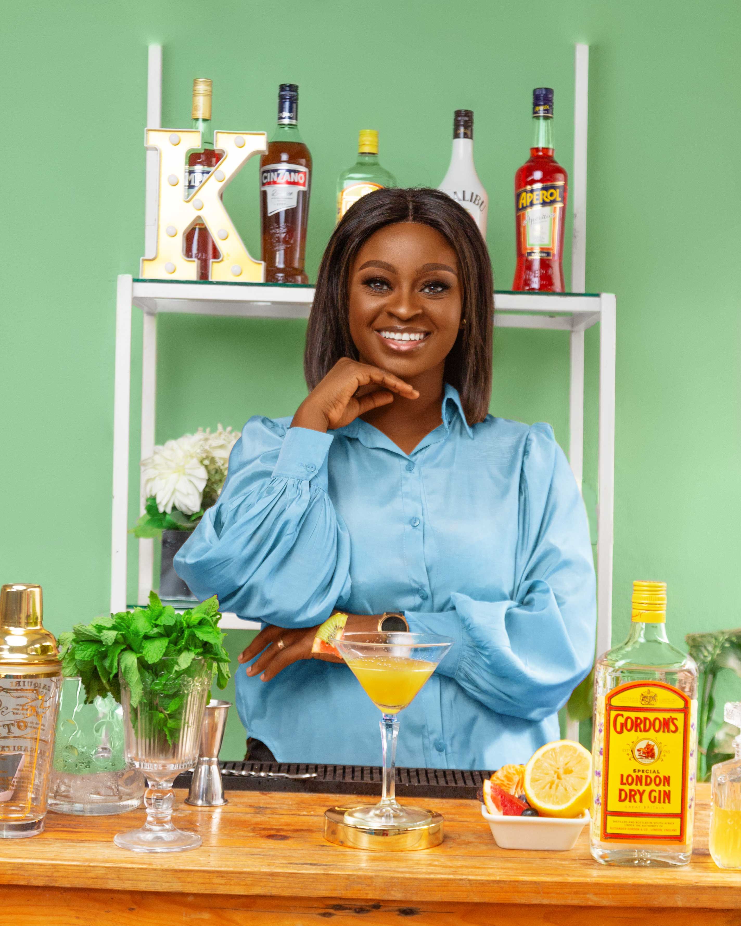 A cheerful bartender in a blue shirt stands behind a bar with various spirits and fresh ingredients in Luanda, Angola.