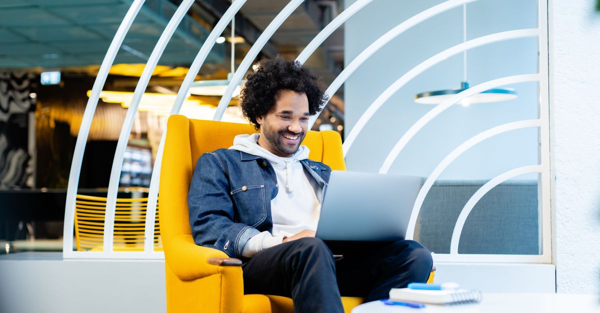 Casual man in denim jacket smiling while working on laptop in a modern office.